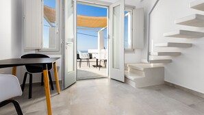 A modern interior with a staircase, a dining area with a table and chairs, and a view through an open door. - SERAPIAS SUITES (Santorini)