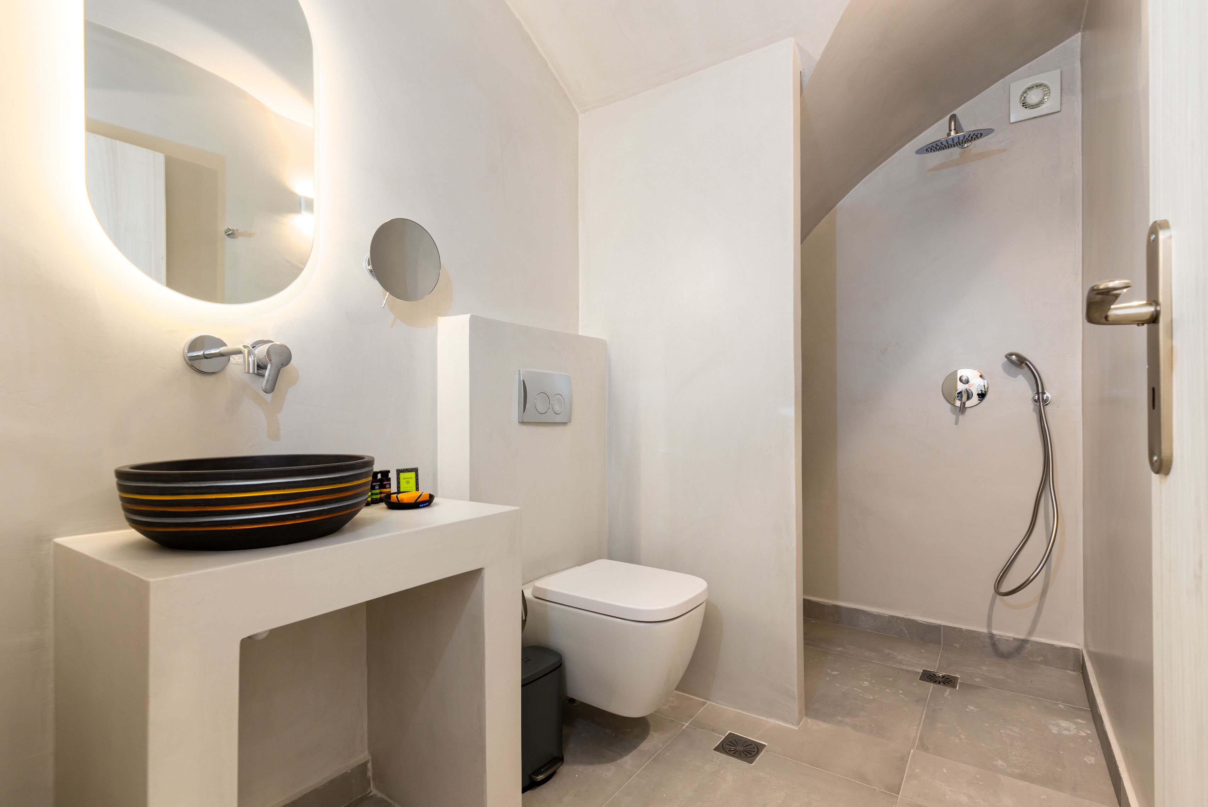 A modern bathroom with a round mirror, a vessel sink, a toilet, and a shower area.