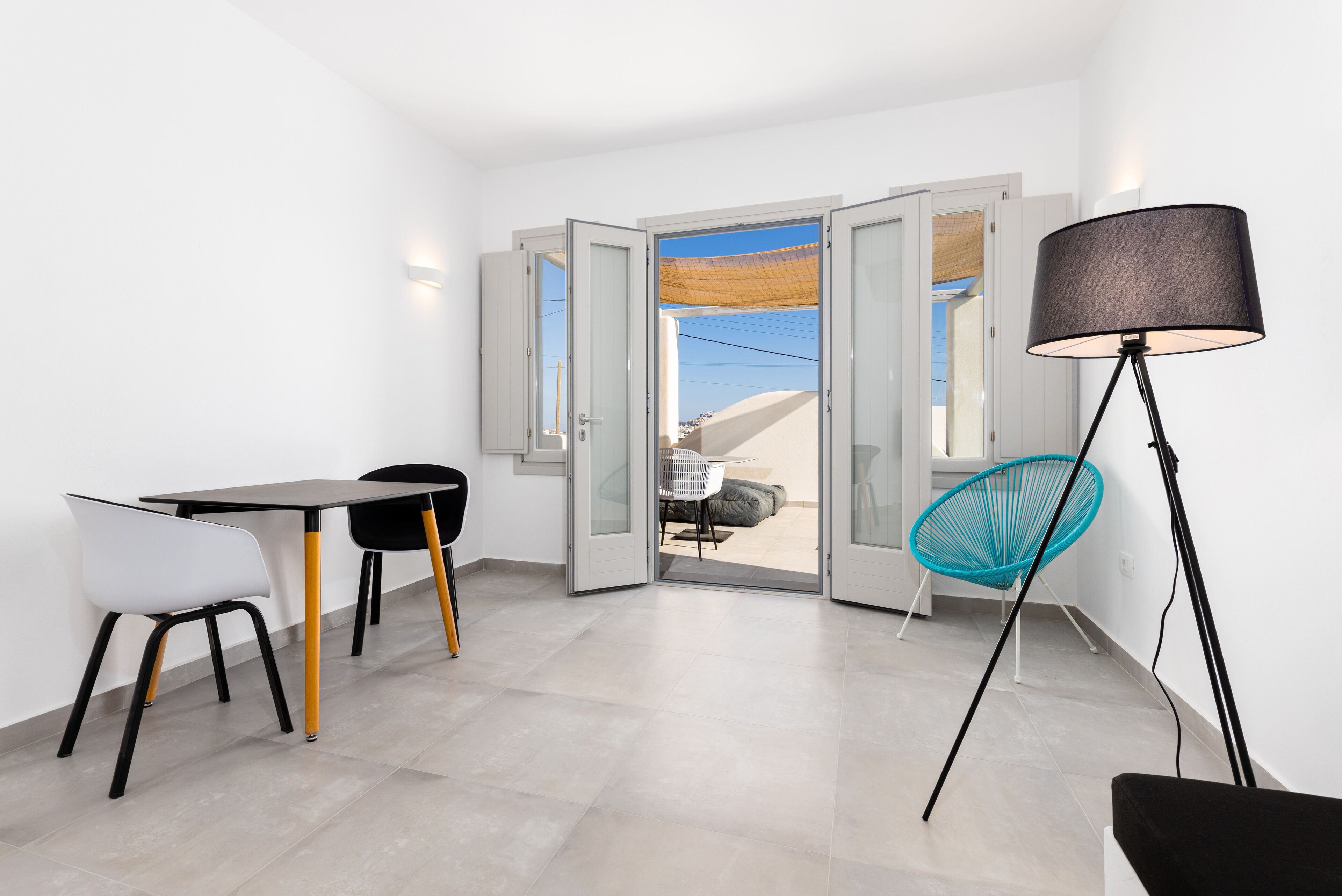 A modern room with a dining table, chairs, a floor lamp, and a view through open doors to an outdoor area.