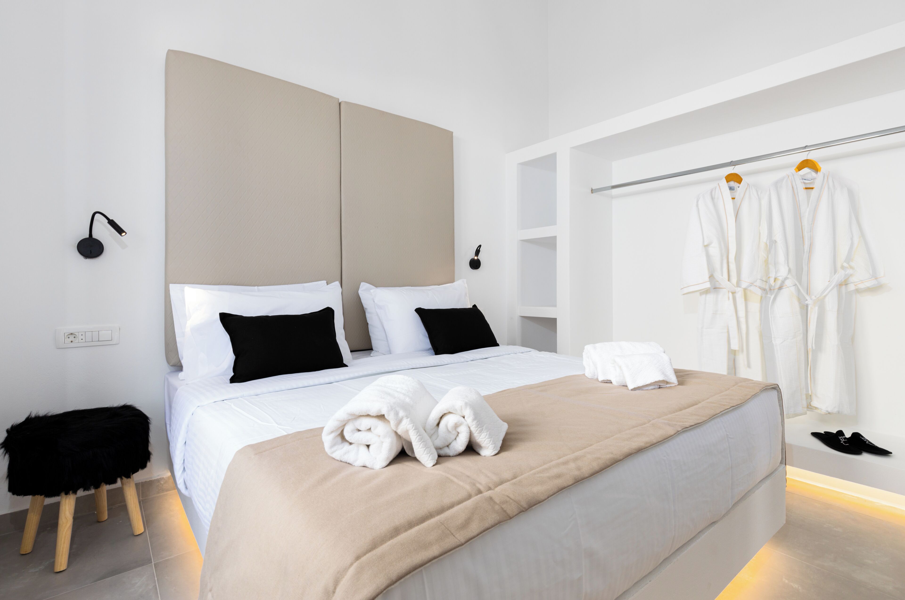 Habitación de hotel moderna con una cama grande, dos almohadas negras, un cabecero color beige, un taburete pequeño, un par de toallas blancas y un perchero con batas y zapatos blancos.
