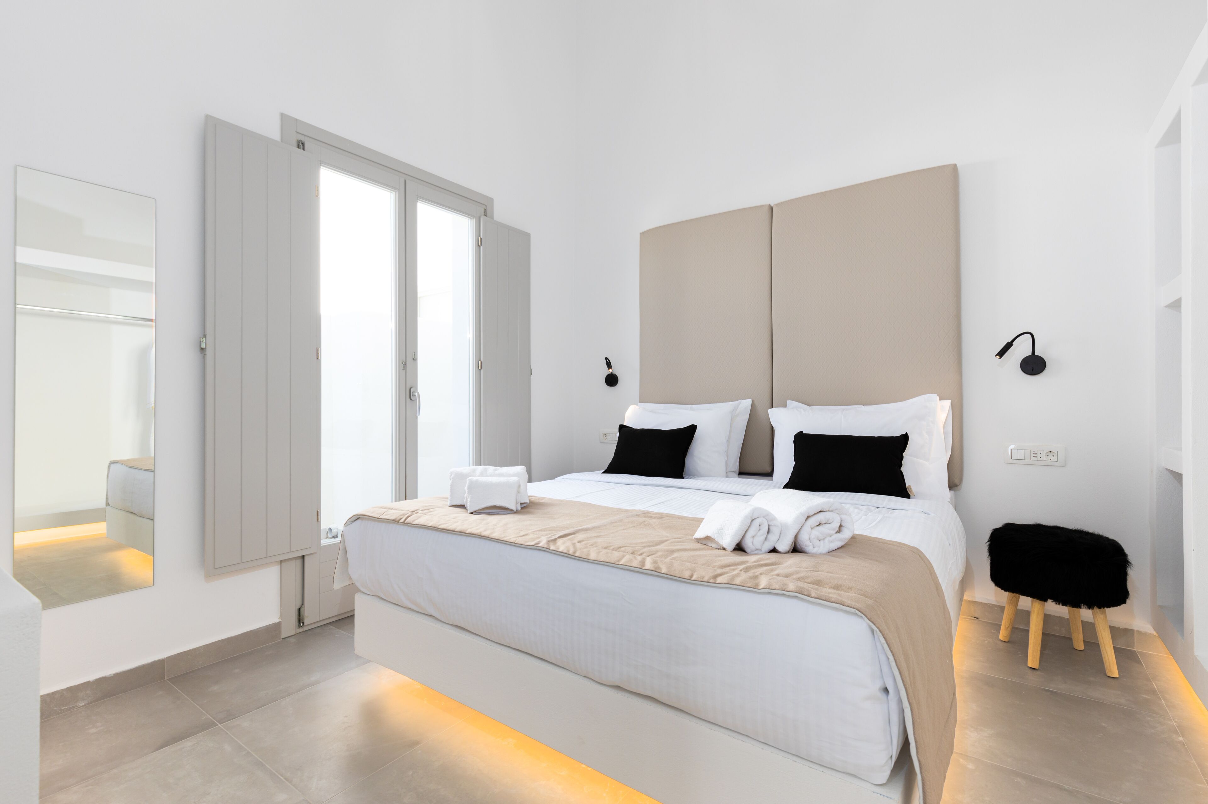 A modern hotel room with a large bed, a bedside table, a mirror, and a small stool.