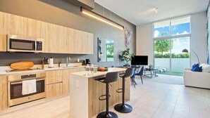 A modern kitchen with a microwave, oven, and bar stools. There is a dining area with a table and chairs, and a large window offering a view of the outdoors. - One Bedroom Condo King Bed With City Views (Miami)