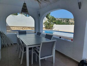 A covered outdoor dining area with a pool view, white furniture, and a stone wall. - Mediterranean Villa with Pool for 12 People (Moraira)