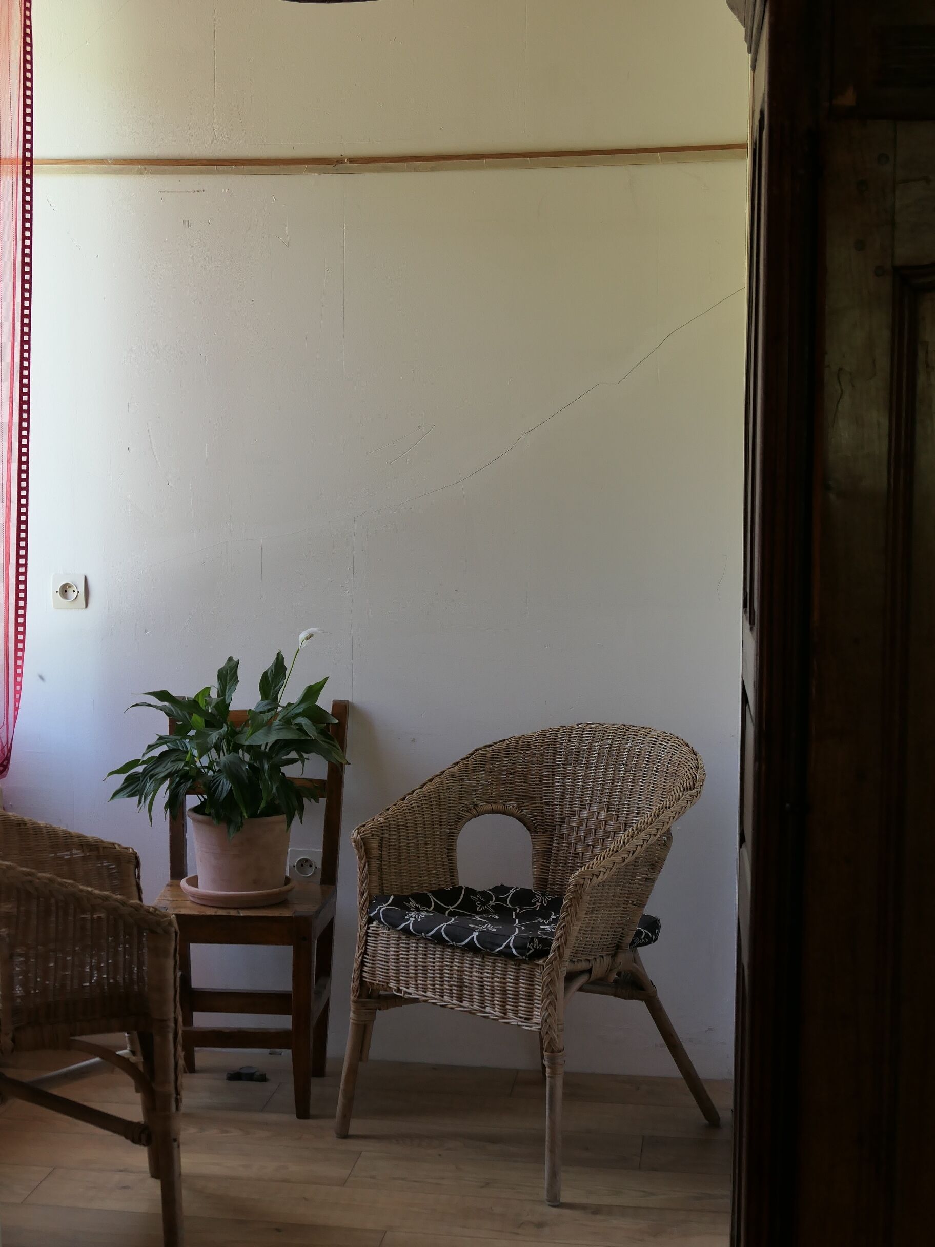 Een rieten stoel met een kussen, een potplant en een rieten tafeltje in een kamer met een houten vloer en witte muren.