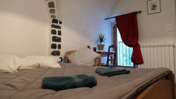 A bedroom with a stone archway, a bed with a beige quilt, a red curtain, a potted plant, and a radiator.
