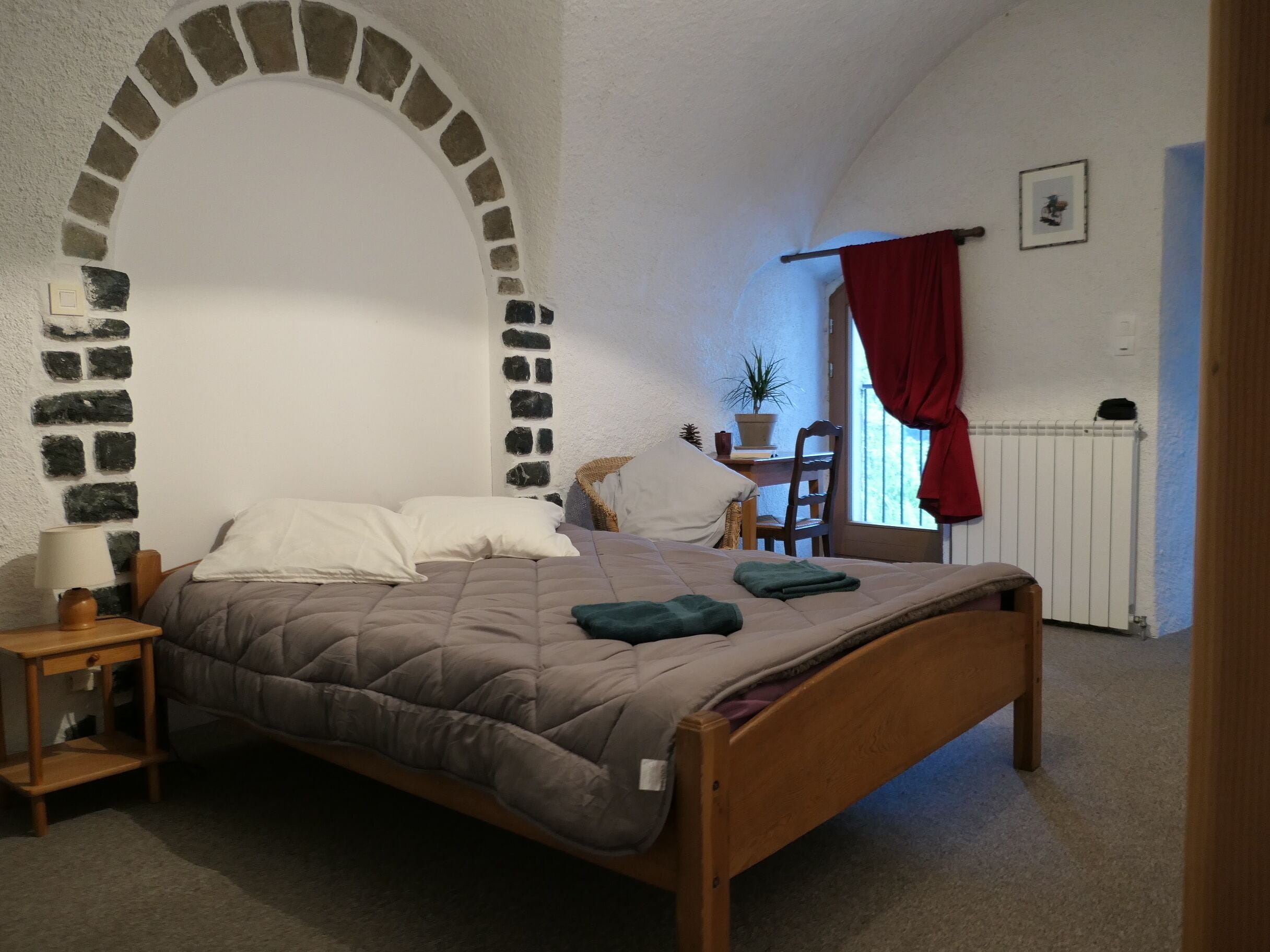 A bedroom with a stone archway, a bed with a quilt, a nightstand, a chair, a desk with a plant, and a radiator.