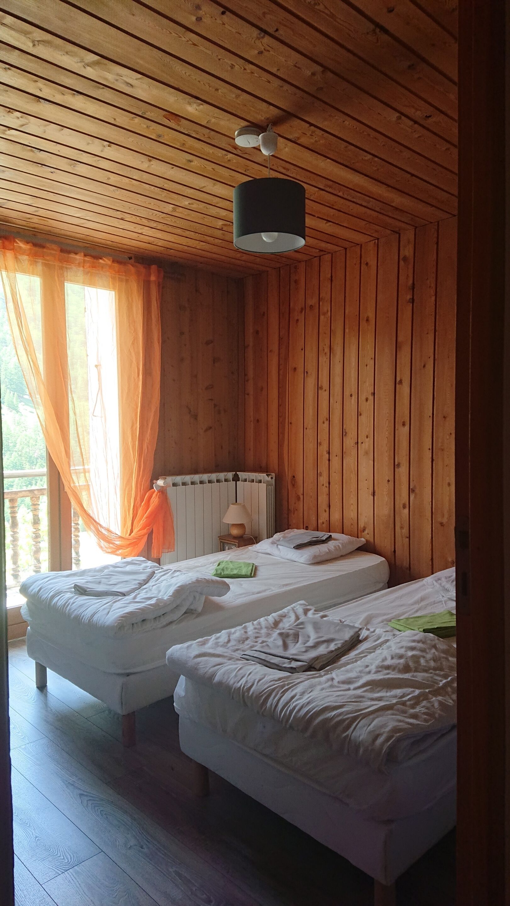 A room with wooden walls and ceiling, two beds with white bedding, a bedside table with a lamp, and a window with orange curtains.