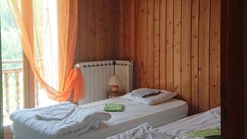 A room with wooden walls and ceiling, two beds with white bedding, a bedside table with a lamp, and a window with orange curtains.