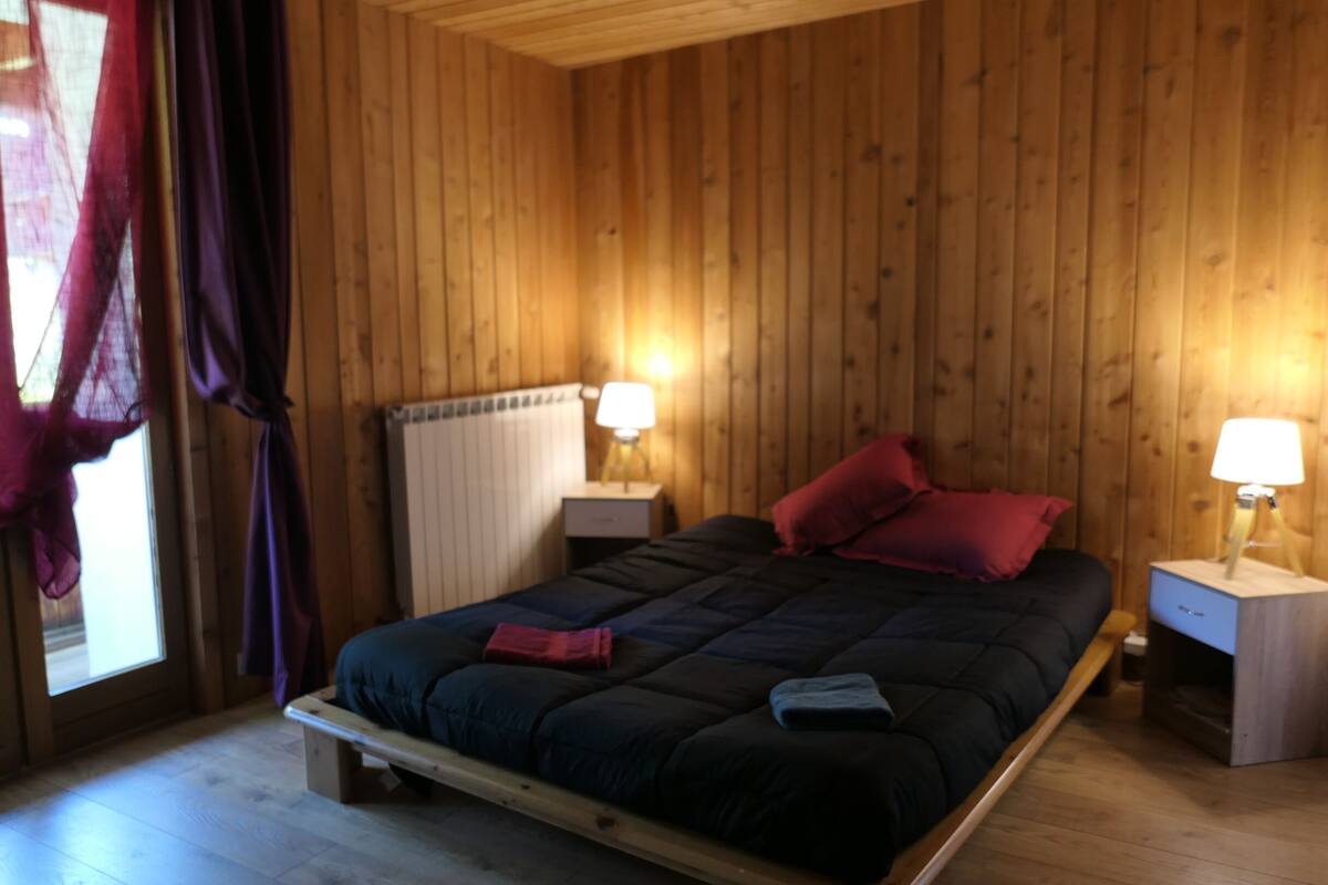Un dormitorio con paneles de madera, una cama, dos mesitas de noche, un radiador y una ventana con cortinas.