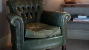 Un sillón de cuero verde con respaldo capitonado, ubicado junto a una mesita de madera con una lámpara y libros.