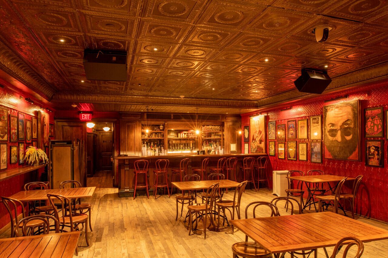 A bar with wooden tables and chairs, a bar counter with bottles, and framed pictures on the walls.
