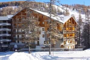 A multi-story wooden building with balconies, surrounded by snow-covered trees and a clear sky.