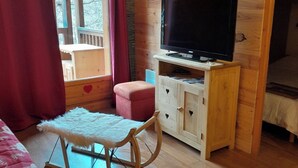 A cozy room with a wooden floor, a television on a wooden cabinet, a rocking chair, and a window with curtains. - L' IDEAL at Ecrin des Neiges. (Vars)