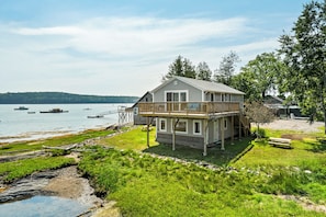 A two-story house with a balcony overlooking a lake, surrounded by greenery and a dock.