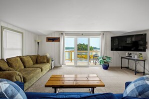 A living room with a sofa, a coffee table, a TV, and a view through a sliding door to an outdoor deck. - Osprey's Nest. Waterfront unit with boat and kayak access. Pet Friendly! (Harpswell)