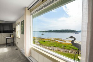 A room with a large window overlooking a body of water, a table with a chair, and a television mounted on the wall. - Osprey's Nest. Waterfront unit with boat and kayak access. Pet Friendly! (Harpswell)