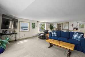 A living room with a blue sofa, a wooden coffee table, a black leather chair, and a view of the sea through a window. - Osprey's Nest. Waterfront unit with boat and kayak access. Pet Friendly! (Harpswell)