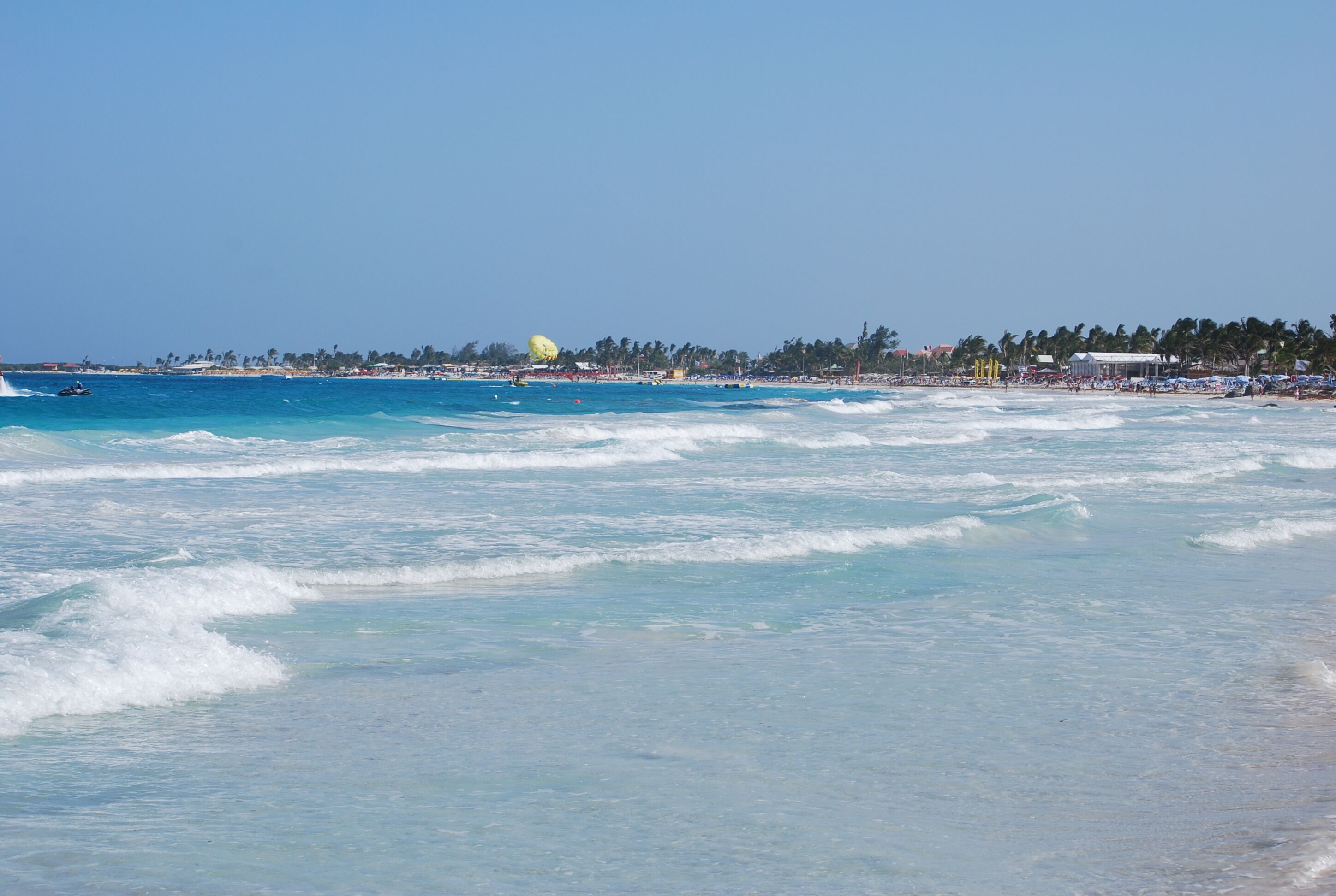 A beach with clear blue water, a sandy shore, and a distant view of a city skyline.