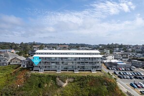 A modern building with multiple floors and a flat roof, surrounded by residential houses and a parking area. - Ocean Front Condo, View of Lighthouse, Walk to Everything, Nye Beach! (Newport)