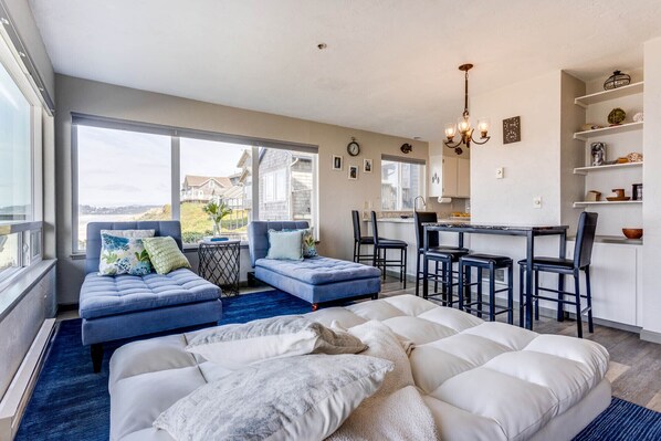 A modern living room with a large bed, blue sofas, and a dining area with bar stools.