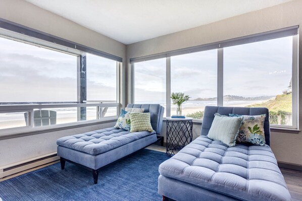 A room with two blue cushioned seating areas, a small round table, and large windows offering a view of the beach and hills. - Ocean Front Condo, View of Lighthouse, Walk to Everything, Nye Beach! (Newport)