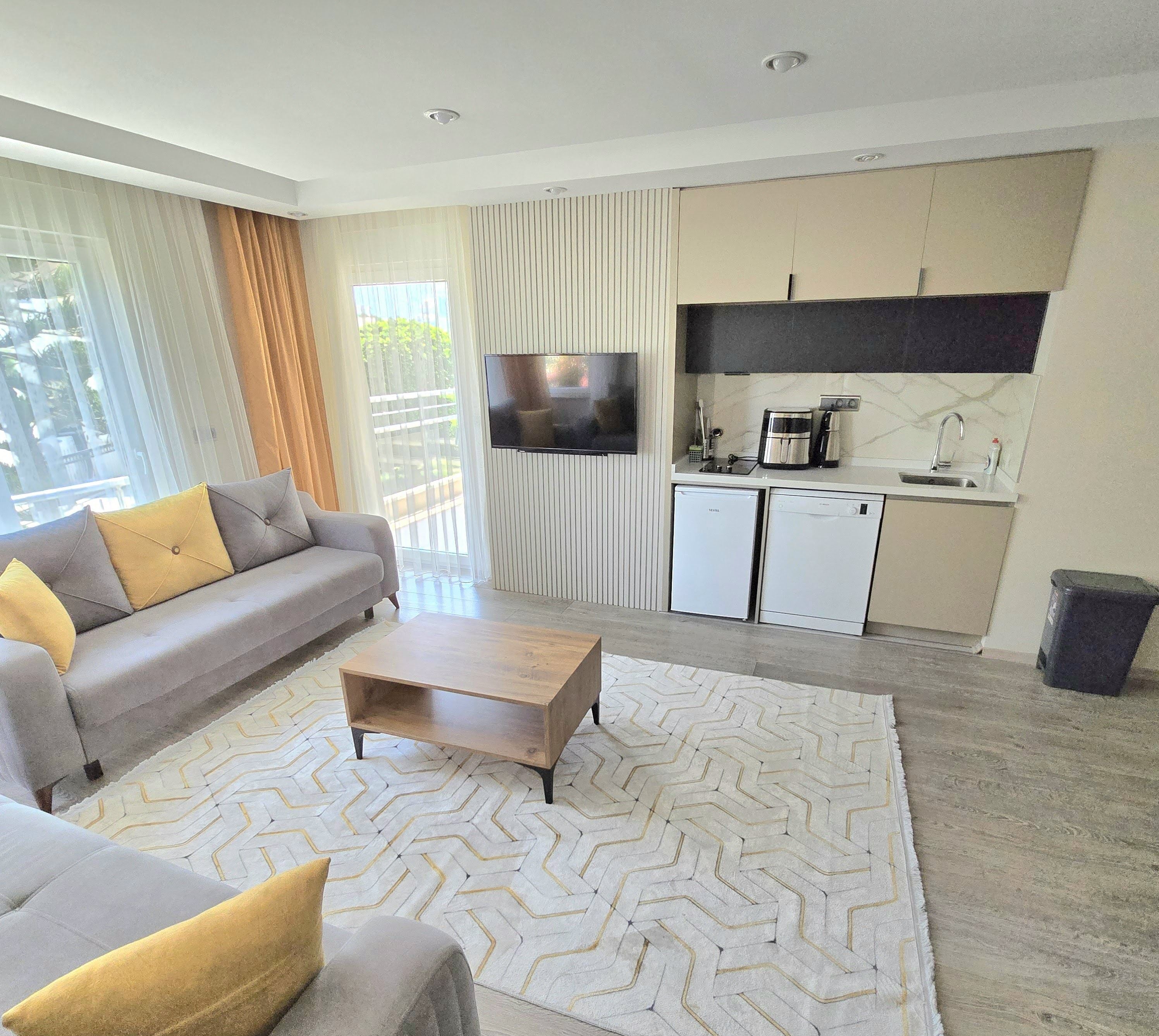 A modern living room with a sofa, coffee table, and a TV. There is a kitchen area with a coffee maker and a sink.