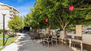An outdoor patio with tables and chairs, surrounded by trees and a building in the background. - River Park Residence Lara (Antalya)