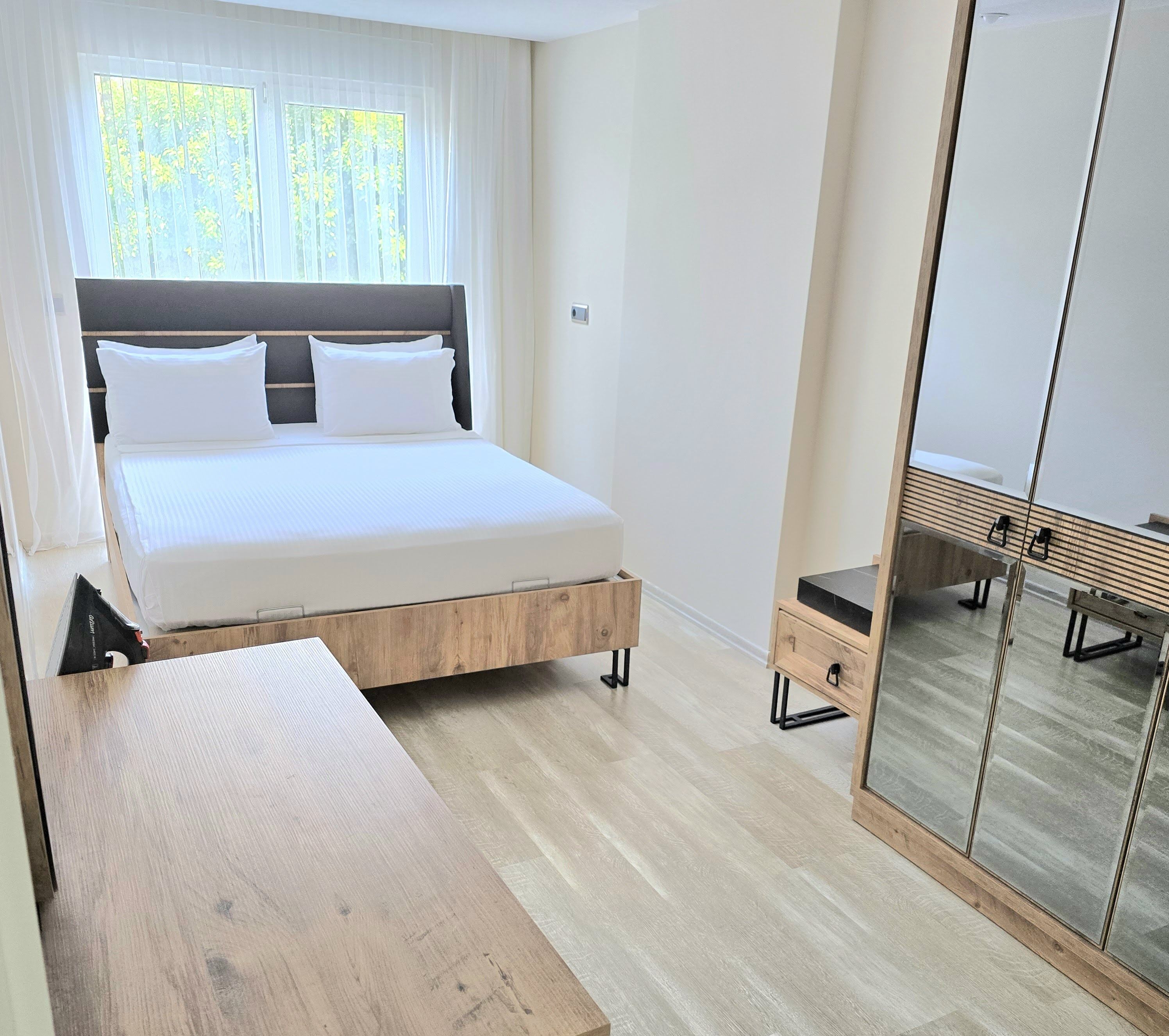 Une chambre à coucher avec un lit, une table de chevet en bois, un miroir et une fenêtre ornée de rideaux légers.