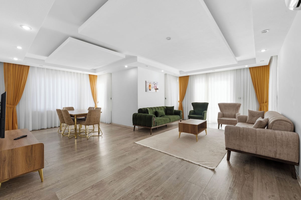 A spacious living room with a wooden floor, a green sofa, a beige sofa, a dining table with chairs, and a TV.