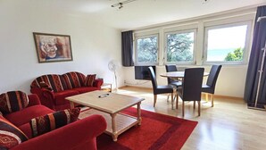A living room with a red sofa, a wooden coffee table, and a dining area with a table and chairs. There is a large window with a view of the outdoors. - Villa Gerster (Wasserburg am Bodensee)