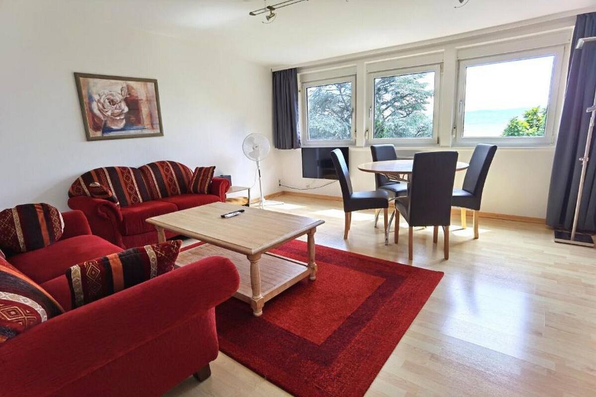 A living room with a red sofa, a wooden coffee table, and a dining area with a table and chairs. There is a large window with a view of the outdoors.