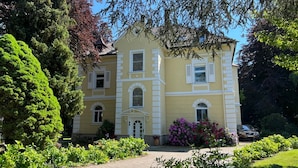 A large, yellow building with white trim, surrounded by lush greenery and flowering plants. - Villa Gerster (Wasserburg am Bodensee)