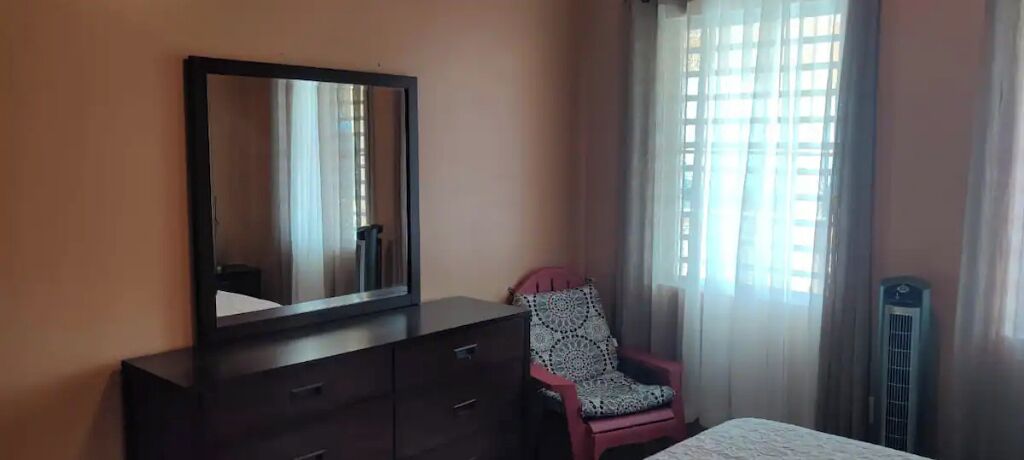 A bedroom with a dark wooden dresser, a chair with a patterned cushion, and a bed with a white cover.