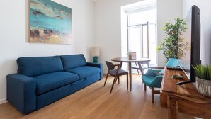 A modern living room with a blue sofa, wooden dining table, chairs, a TV, and a painting on the wall.