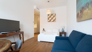 A modern living room with a flat-screen TV, wooden coffee table, blue sofa, and a bed with white bedding.