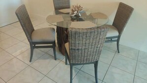 Uma mesa de vidro redonda com quatro cadeiras de vime, uma cesta pequena de fibra natural e um vaso com flores secas.
