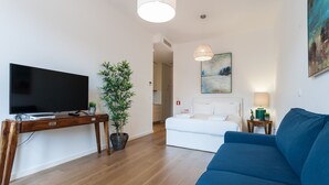 A modern living room with a flat-screen TV, wooden console table, potted plant, blue sofa, white bed, and a painting on the wall. - FLH Restauradores Modern Studio II (Lisbon)