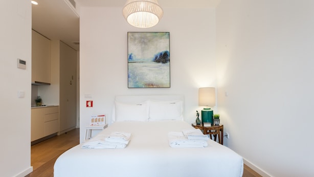 A hotel room with a large bed, a painting on the wall, a bedside table with a lamp, and a view into the kitchen. - FLH Restauradores Modern Studio II (Lisbon)