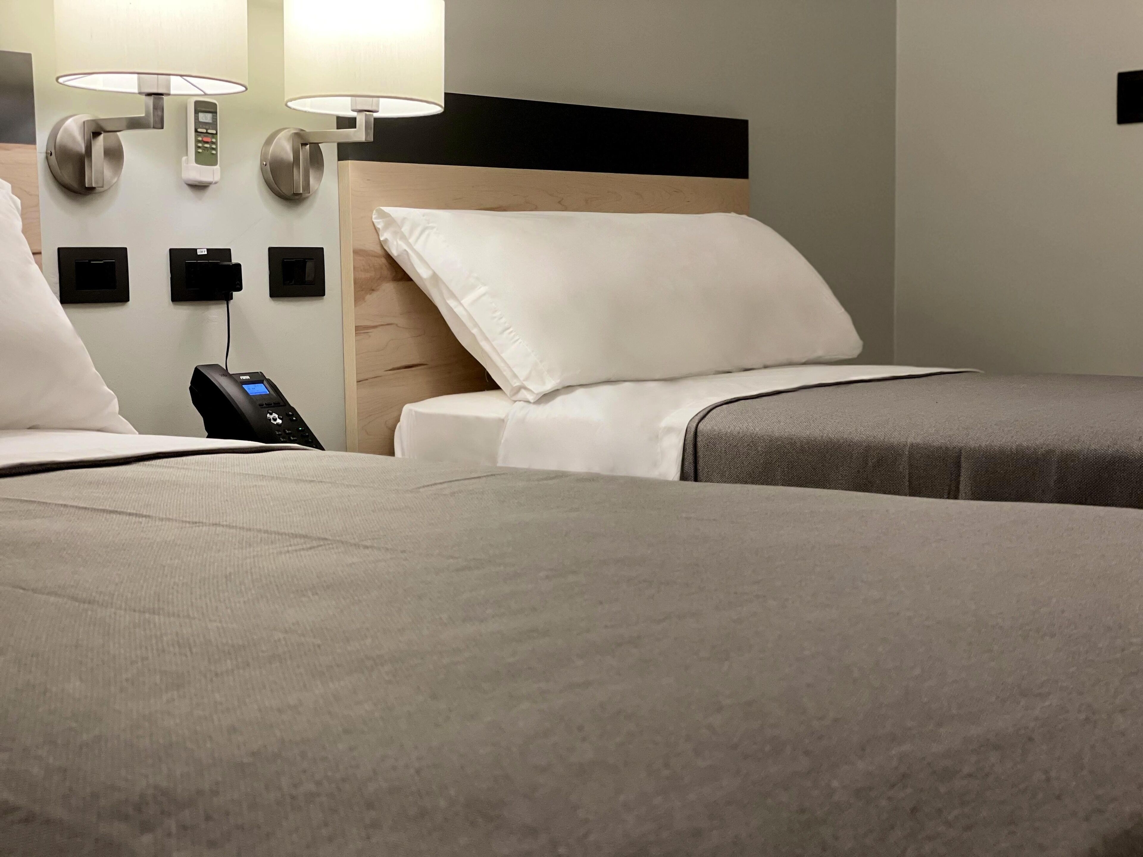 A hotel room with two beds, a bedside table with a phone and a lamp, and a wall-mounted control panel.
