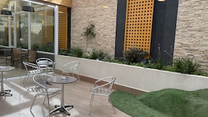 Un espace extérieur aménagé avec des chaises et des tables en métal, un mur en pierre et un petit jardin avec du gazon synthétique et des plantes.