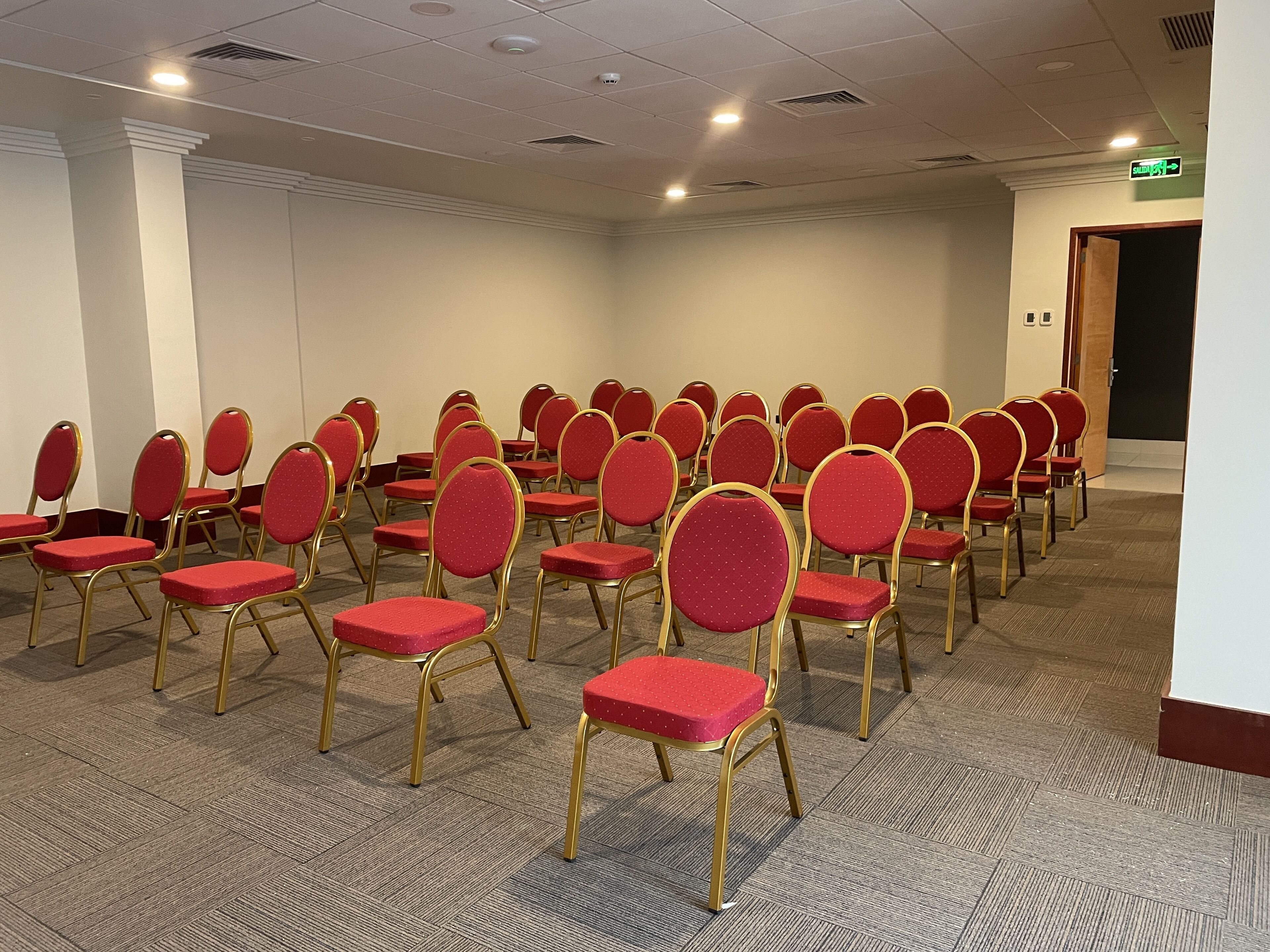 A conference room with rows of red chairs and gold frames, carpeted floor, white walls, and ceiling lights.