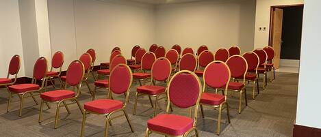 A conference room with rows of red chairs and gold frames, carpeted floor, white walls, and ceiling lights.