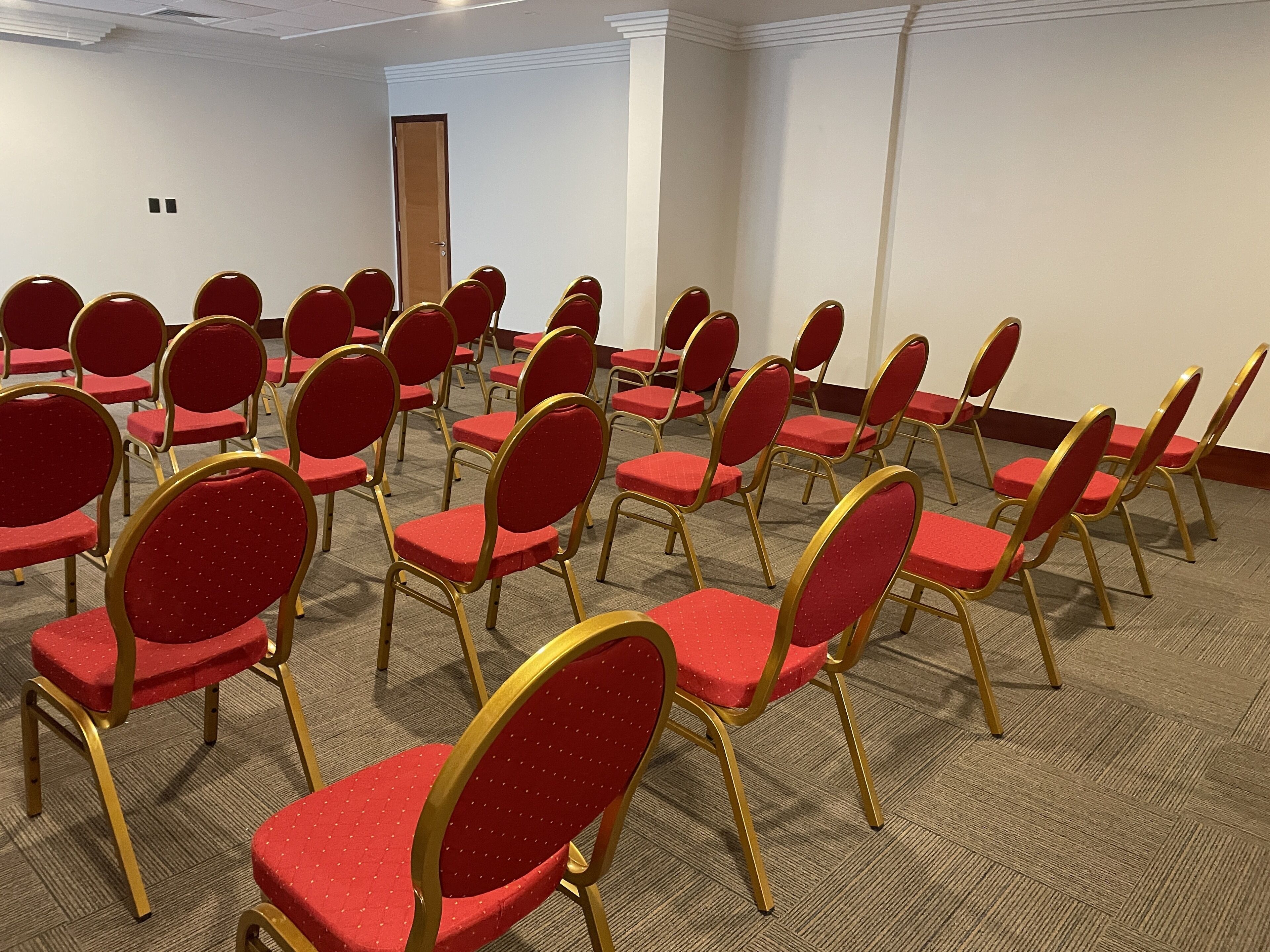 Un salón de conferencias con filas de sillas rojas y marcos dorados, piso alfombrado y una puerta al fondo.