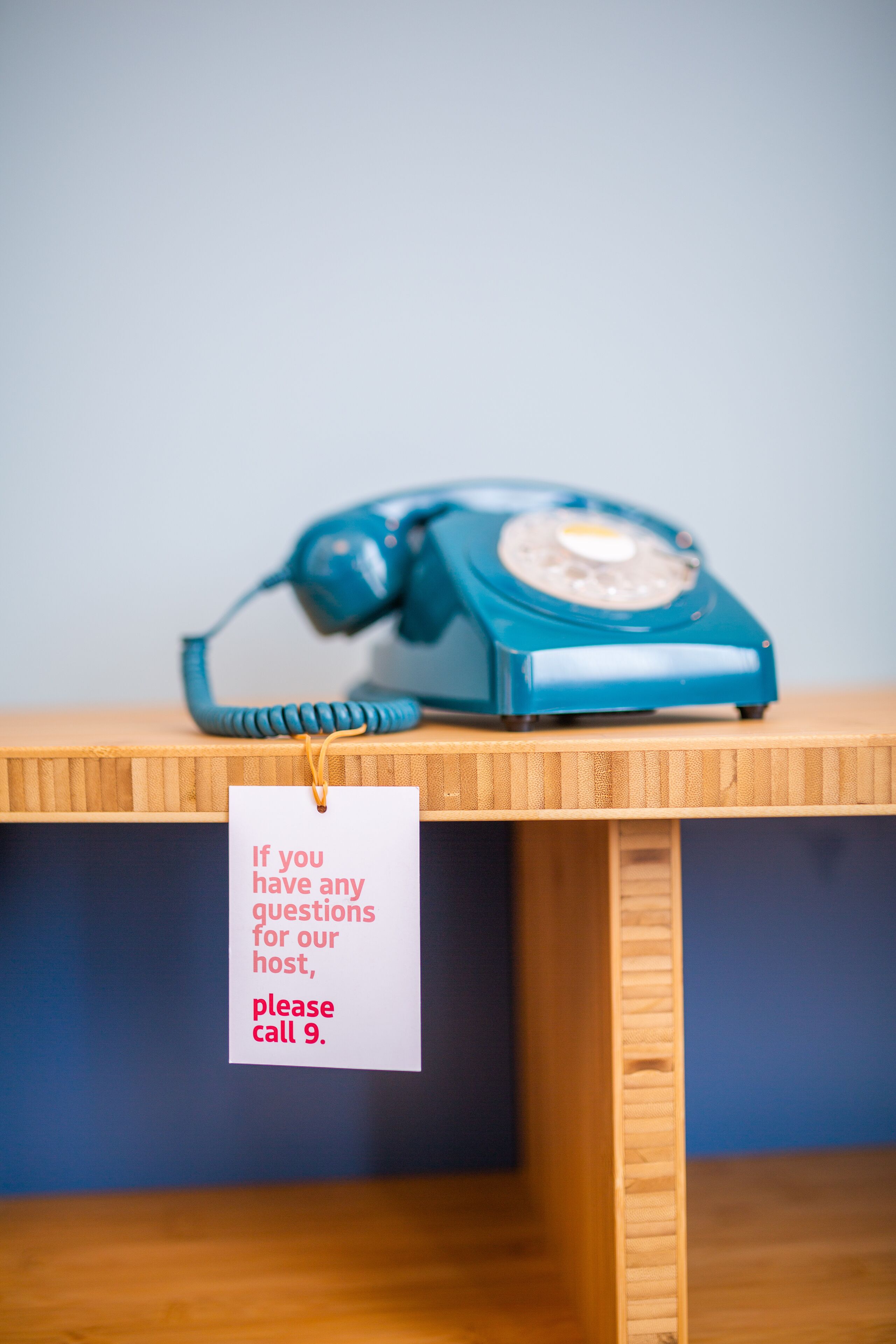 A blue rotary phone on a wooden table with a tag that reads "If you have any questions for our host, please call 9."