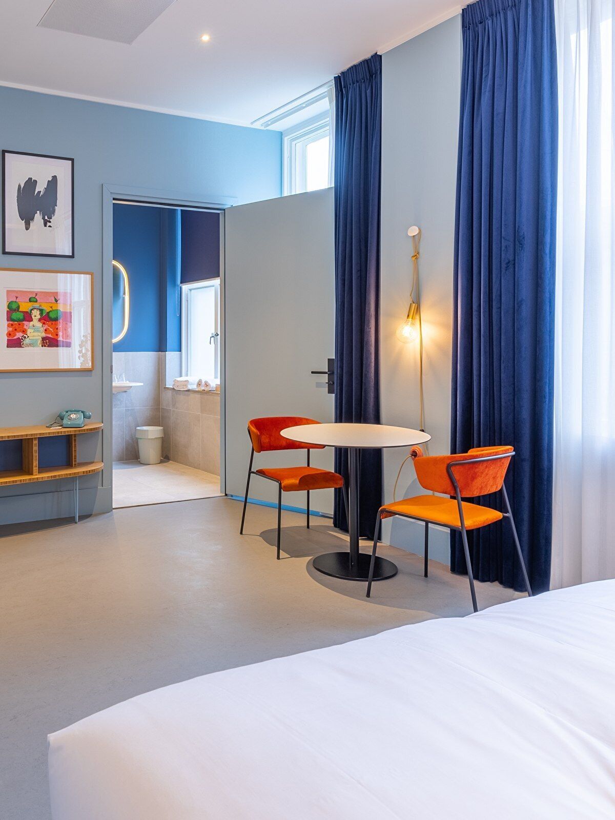 A modern hotel room with a bed, a round table with two orange chairs, a small desk with a telephone, and a bathroom visible through an open door.