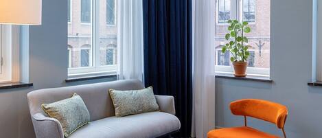 A modern living room with a grey sofa, an orange chair, a small wooden table, and a potted plant by a window with white curtains.