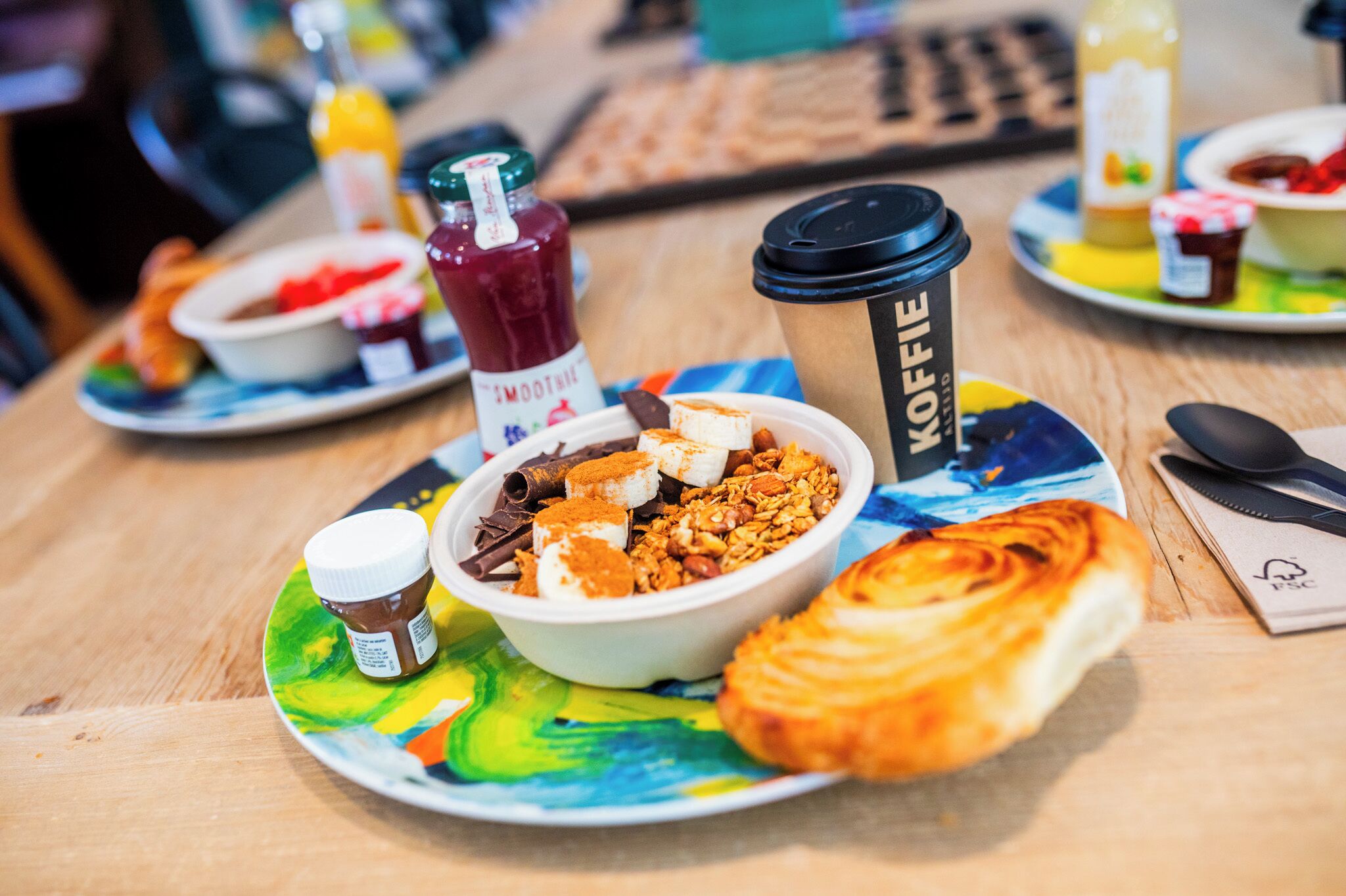A breakfast setting with a bowl of cereal, a croissant, a coffee cup, and a bottle of smoothie.