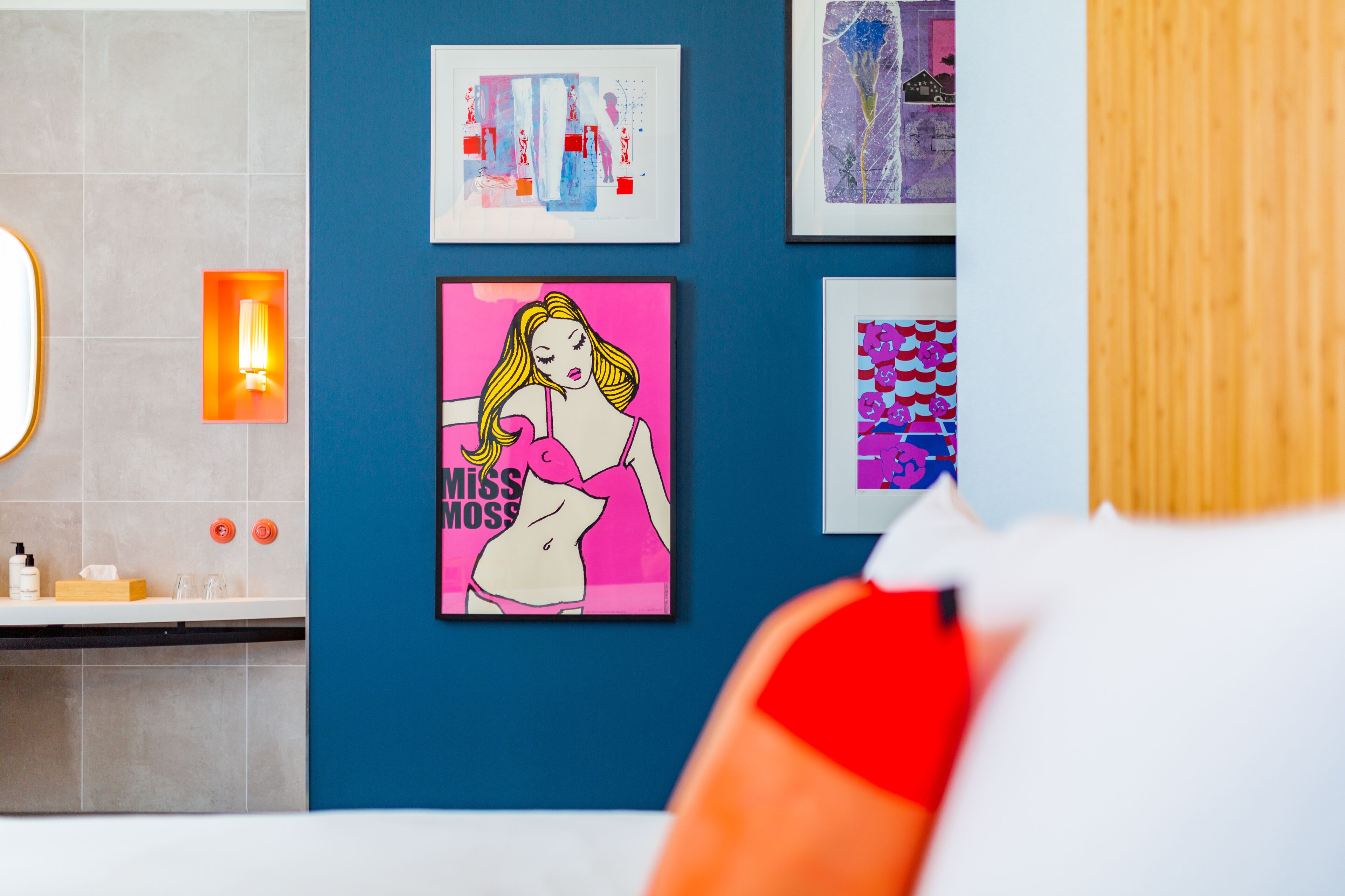 Un baño con pared azul, un lavabo y un espejo. Hay obras enmarcadas en la pared, incluyendo un cartel rosa con una mujer y el texto 