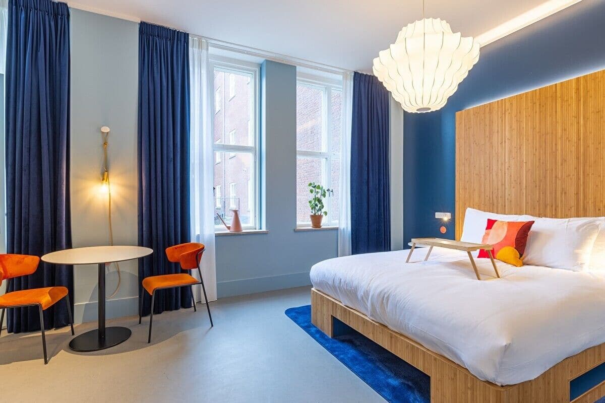 A modern hotel room with a large bed, a round table with orange chairs, and a view of the city through large windows.
