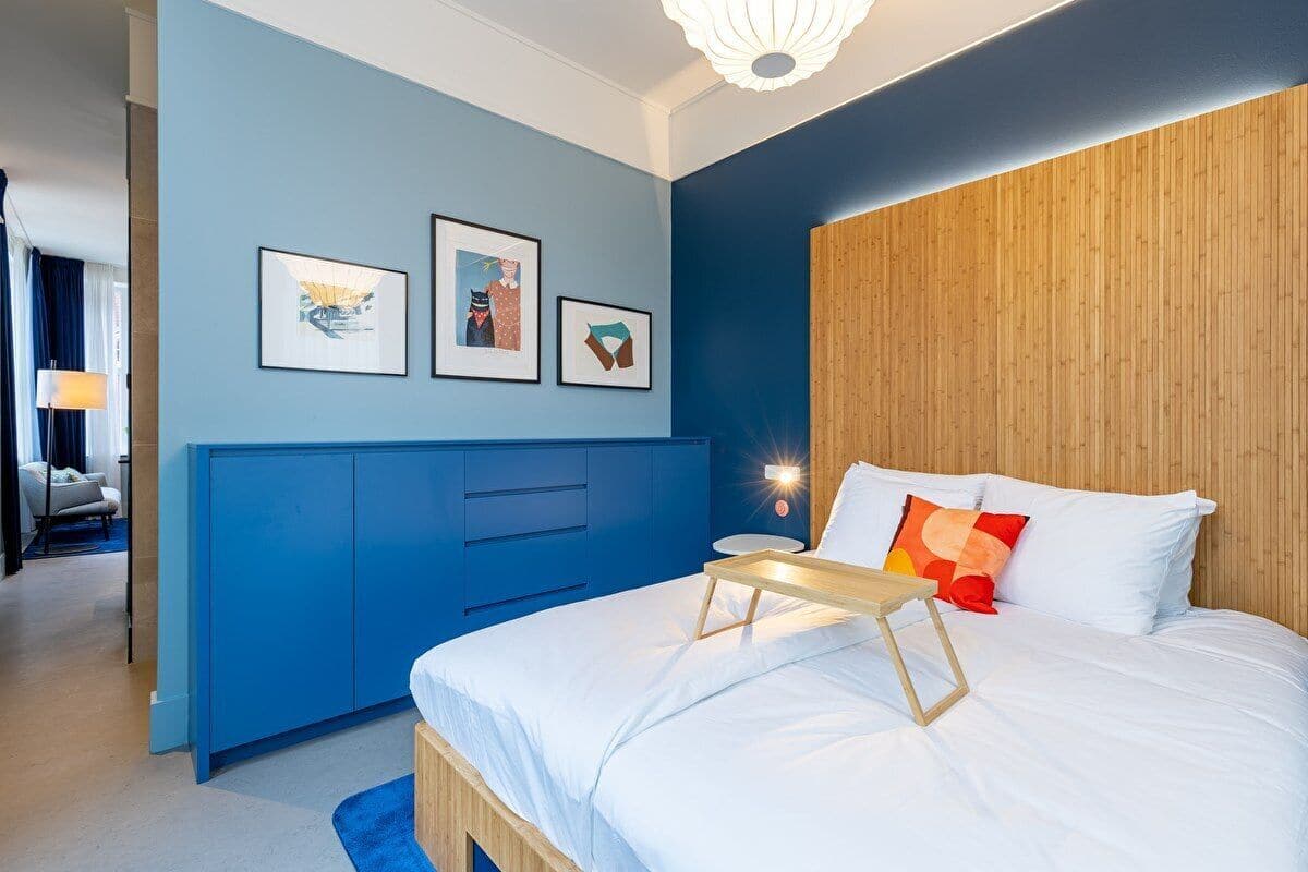 A modern hotel room with a blue accent wall, a wooden headboard, a bed with white bedding, and a small bedside table.